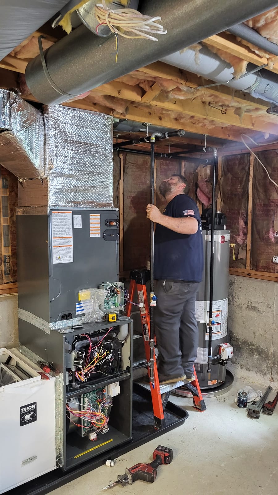 Plumber working on HVAC system installation in basement setting, with visible furnace unit, tools on the floor, and insulated ductwork in the background.