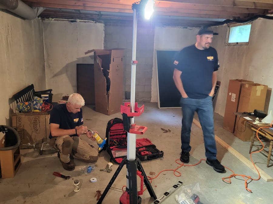 Two workers in a basement surrounded by tools and equipment.