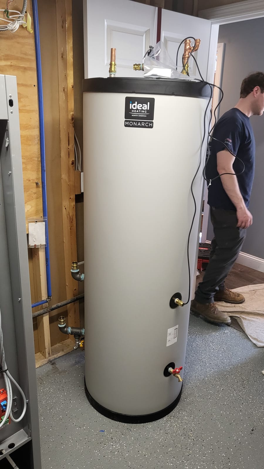A plumber installs an Ideal Heating water heater in a basement with visible piping and electrical connections.
