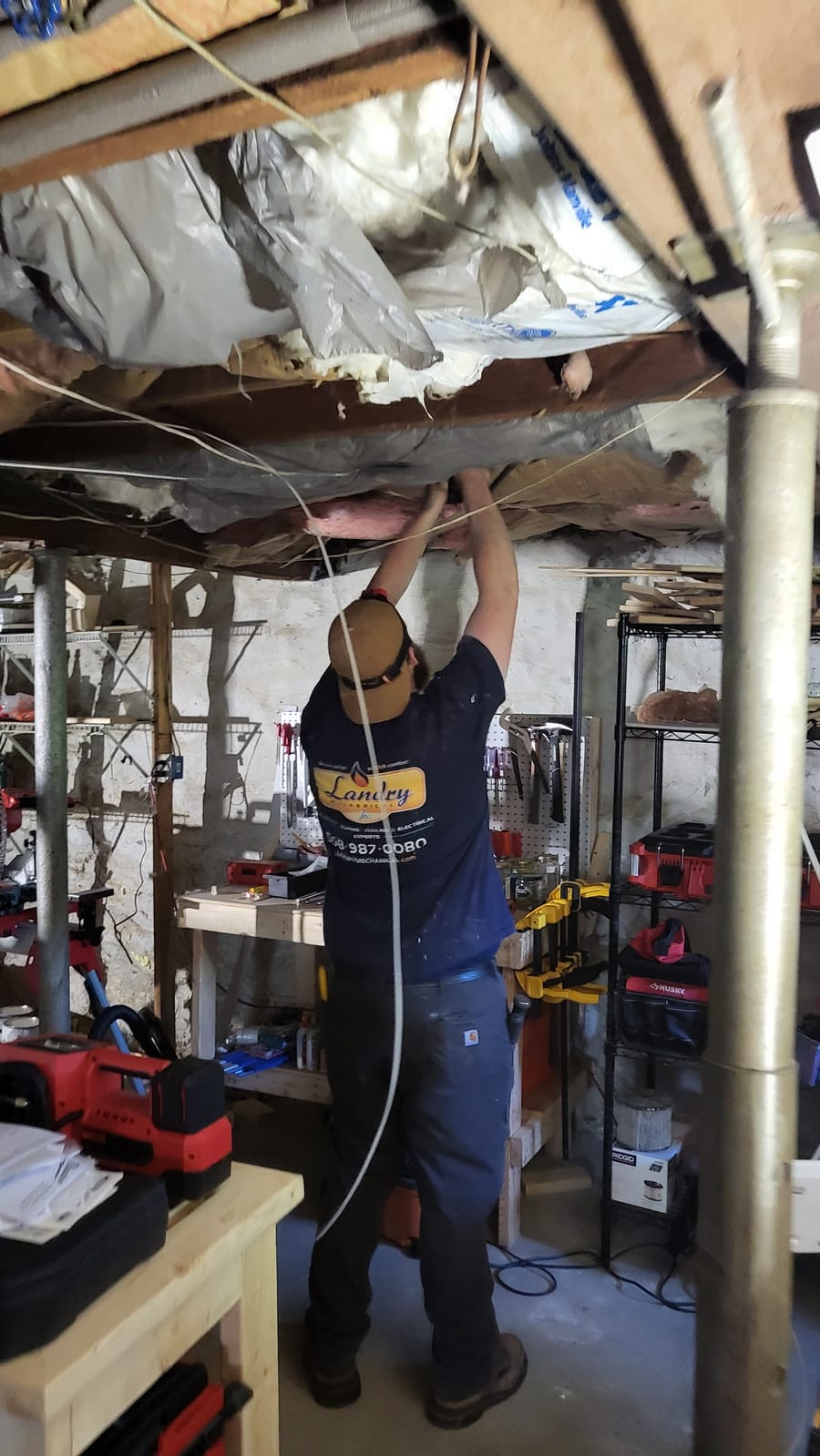 Plumber working on ceiling pipes in a basement workshop, surrounded by tools and equipment.