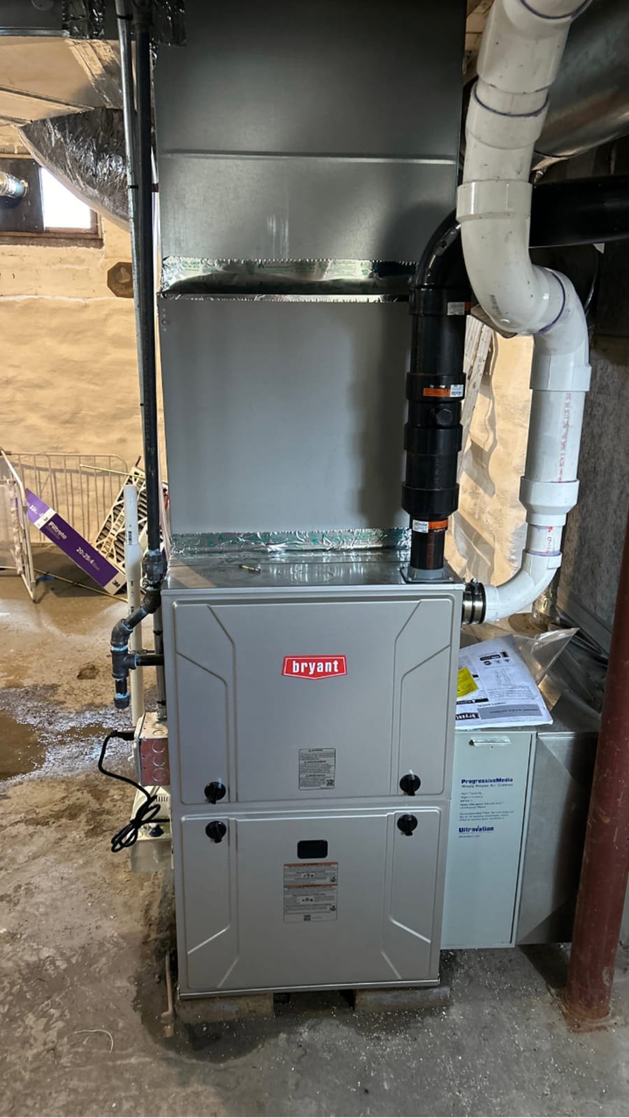 A Bryant furnace setup in a basement, showing connected piping and ventilation, with installation materials and a partially visible window.