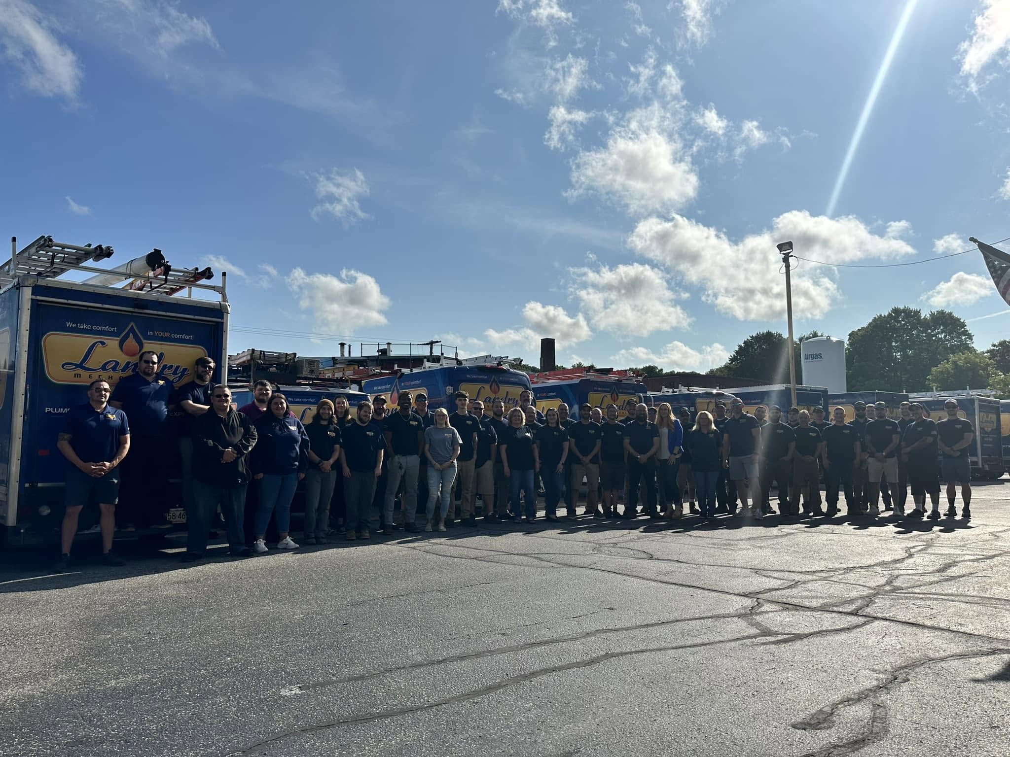 Group of employees posing for a team photo in a parking lot beside blue delivery/service trucks on a sunny day.