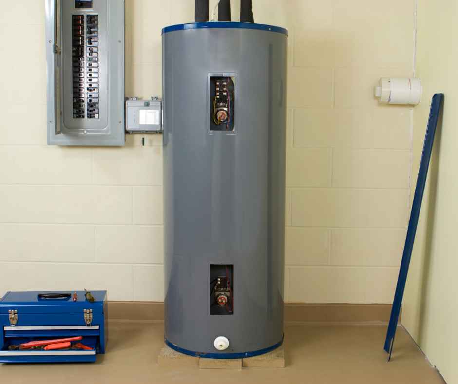 Tall gray vertical water heater in a utility room with two access panels opened to reveal valves and wiring, next to an electrical panel on the wall and a blue toolbox on the floor.